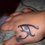 A small, blue Ankh with an eye in the center, tattooed on the back of a hand. The style is traditional with bold lines.