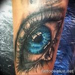 A detailed black and grey tattoo of a realistic blue eye with intricate eyelashes, covering a portion of the forearm. A captivating example of realism tattoo art.