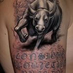 Black and grey tattoo of a charging bull with ornate script below, placed on the side. A dynamic and detailed tattoo design.