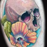 Skull tattoo with a vibrant orange hibiscus flower growing from it, a colorful and symbolic memento mori design.