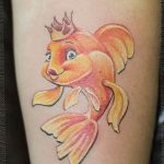 Cartoon-style koi fish tattoo with a crown on the forearm, featuring bright colors, bold outlines, and a playful aesthetic.