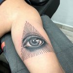 Black and grey tattoo of a realistic eye surrounded by radiating lines, creating a sunburst effect on the upper arm.