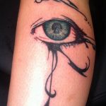Black and teal tattoo of a realistic weeping eye with stylized tribal swirls on the forearm, showcasing detailed shading and emotional expression.