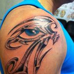 Black and grey tattoo of the Eye of Horus with tribal-inspired design elements on the shoulder, featuring a blue eye.