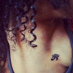 Small black ink tattoo of the Egyptian Eye of Horus on the upper chest, near the collarbone, in a minimalist style.