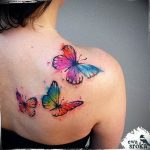 Vibrant watercolor butterfly tattoos on the shoulder, featuring splashes of pink, blue, and yellow, artistic and flowing style.