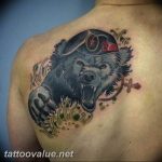 Colorful tattoo on the shoulder featuring a roaring bear wearing a military cap, with floral elements and a cross, symbolizing strength and affiliation.