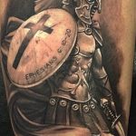 A detailed black and grey tattoo on the upper arm depicts a Spartan warrior in full armor, with a shield bearing a cross and the inscription 'Ephesians 6:10-20'.