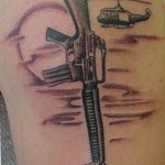 Black and grey realism tattoo featuring a helmet, rifle, and boots, with a helicopter in the background, honoring a fallen soldier.