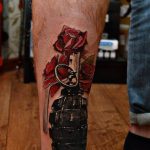 Realistic tattoo of a grenade with red roses blooming from the top, on the calf, showcasing detailed shading and color.