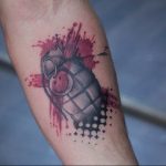 Black and red watercolor style tattoo of a grenade with red splatter effects and black dot work, on the forearm, representing chaos and impact.