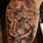 Detailed black and grey tattoo of a dove perched above a rose, on the upper arm. A romantic tattoo design combining symbols of peace and love.