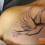 A minimalist black line-art tattoo of a dove in flight, featuring flowing lines and a simple design, placed on the shoulder.