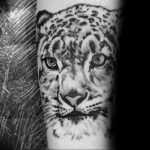 Detailed black and grey realism tattoo of a snow leopard's face, focusing on the eyes and fur texture, placed on the forearm.