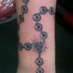 A large black and grey tattoo of a rosary wrapping around the entire forearm and hand, featuring detailed beads and a crucifix. A full sleeve-style tattoo.