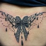 Black ink tattoo featuring a detailed bow with lace and vine-like elements across the lower back. A decorative and feminine tattoo style.