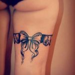 A detailed black ink tattoo of a lace bow encircles the thigh, creating a garter-like effect with a sophisticated aesthetic.