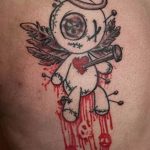 Dark tattoo of a voodoo doll with angel wings, a halo, and dripping blood, on the back. A gothic and symbolic tattoo design.