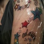 A colorful tattoo on the upper arm featuring various starbursts and the name 'adash' in script, with a playful and vibrant aesthetic.