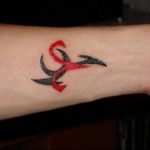 A small Aries symbol tattoo on the wrist, using a combination of red and black ink for a striking two-tone effect.