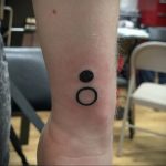 Small black tattoo featuring a filled circle above an open circle, representing binary code on the wrist. Minimalist, geometric style.