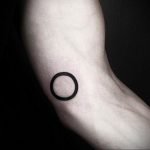 Small, minimalist black circle tattoo on the inner bicep, showcasing a clean and understated design.