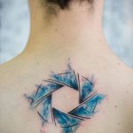 A watercolor-style tattoo of a camera aperture on the upper back, featuring blue and grey washes. The design is artistic and fluid.