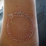 White ink tattoo of a dragon-like serpent forming a circle, with a textured scale pattern, on the forearm. A unique and subtle symbolic tattoo.