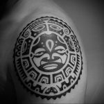 A black Polynesian tattoo on a shoulder, featuring a stylized face, traditional patterns, bold lines, cultural ink, tribal art.