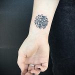 Small, detailed floral tattoo on the wrist, circular arrangement, black ink, representing nature, growth, and beauty.