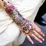 Detailed floral tattoo sleeve on a hand, featuring red and green blooms with black linework, in a delicate illustrative style.