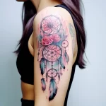 Watercolor dreamcatcher tattoo with roses and feathers, adorning the shoulder and upper arm in a delicate style.