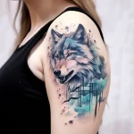 Watercolor tattoo of a wolf's head with piercing eyes and abstract blue splashes on the shoulder. Features a realistic portrait style.