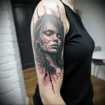 Black and grey tattoo of a woman's face with cracked skin and a single rose, on the upper arm. Dramatic shading and a melancholic aesthetic.