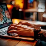 A close-up of a hand and forearm with a detailed tattoo, while the person is typing on a laptop. The tattoo features floral and possibly animalistic elements.