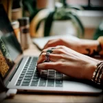 A close-up of a hand with a small, dark tattoo on the back of the hand, while the person is typing on a laptop.