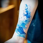 Realistic blue wave tattoo wrapping around a forearm, showcasing fluid motion and detailed water effects in a watercolor style.