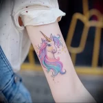 A vibrant watercolor-style unicorn head tattoo graces the forearm, featuring a rainbow-colored mane and delicate details.