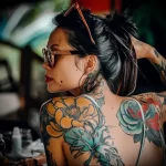 A woman viewed from behind, showcasing a large, colorful floral tattoo covering her back and shoulder. She is wearing sunglasses and earrings.