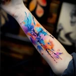 A watercolor tattoo on the forearm featuring abstract shapes and flowing colors in shades of blue, purple, and orange.
