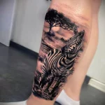 Realistic black and grey zebra tattoo within a savannah landscape on the calf, showcasing detailed animal portraiture and scenic background.