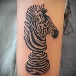 Black ink zebra head stylized as a chess knight tattoo on the arm. A creative and symbolic tattoo design.
