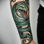 Colorful tattoo of a green dragon wrapping around a man's forearm. Detailed scales, sharp claws, and an open mouth create a fierce and dynamic image.