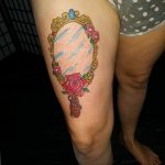 A colorful tattoo on the thigh featuring a vintage-style mirror reflecting a rose, framed with delicate floral accents. The style is illustrative and traditional.